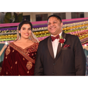 ​Dr. Michael Thomas smiling in a black tuxedo alongside Dr. Praisey Thomas (née Yesudas) wearing a traditional embroidered maroon velvet lehenga at their wedding reception at Cross Ground in Itarsi, Madhya Pradesh, India.